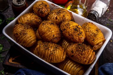 Homemade hasselback potatoes it baking tray. Baked hasselback potatoes with cheese, garlic and herbs, on kitchen table with ingredient for cooking