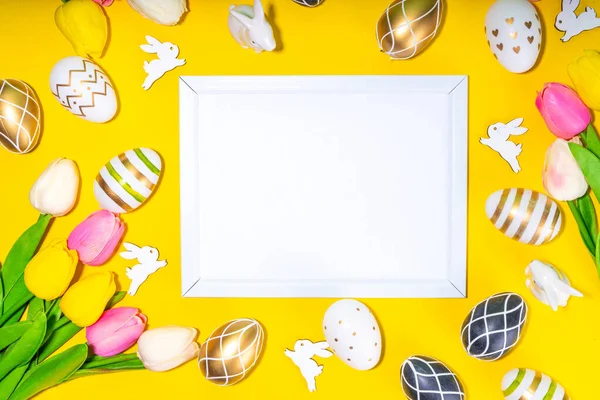 Bright Easter greeting card background. Trendy colored black, golden, stripe painted Easter eggs with white bunny rabbits decor, tulip flower on high-colored yellow background, flat lay copy space