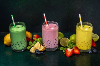 Üç renkli yaz köpüklü çay seti, parlak kremalı inci çayı ya da latte tapyoka içecekleri, pembe dut, sarı narenciye, yeşil nane, tapyoka topları ve ezilmiş buz, koyu arkaplanda.