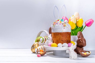 Şekerli Paskalya pastası, renkli yumurta sepeti, seramik tavşanlar ve beyaz mutfak masasında bahar çiçeği, kopyalama alanı. Mutlu Paskalya Tatilleri tebrik kartı arkaplan kopyalama alanı