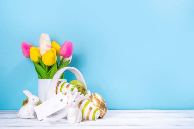 Paskalya Bayramı tebrik kartı geçmişi. Bahar çiçekleri buketi, renkli yeşil, tavşanlı Paskalya yumurtası sepeti ve beyaz ahşap ve mavi arkaplan üzerinde Mutlu Paskalyalar etiketi