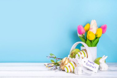 Paskalya Bayramı tebrik kartı geçmişi. Bahar çiçekleri buketi, renkli yeşil, tavşanlı Paskalya yumurtası sepeti ve beyaz ahşap ve mavi arkaplan üzerinde Mutlu Paskalyalar etiketi