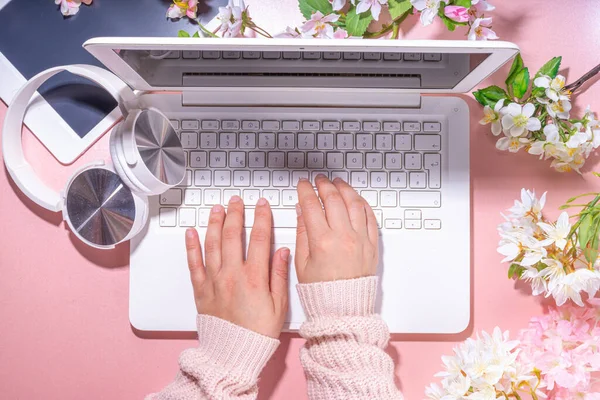 Spring office workplace, blogging flat lay background. White laptop, with headphones, tablet, spring flowers bouquet on pink pearl background, girl\'s hands typing on a laptop.