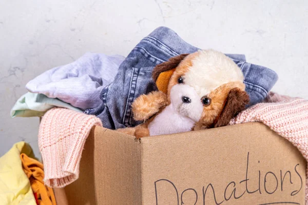 stock image Donation box with clothes, toys and food on white table background. Cardboard donation box full with toys, female and child clothes, foods. Social care, volunteering, charity concept copy space