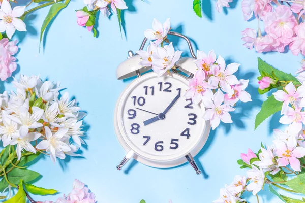 Spring Time Change background. Beautiful fresh spring tree flowers with alarm clock on light blue sunny background top view copy space