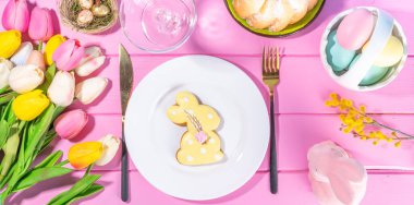 Şirin Paskalya sofrası, bahar tatili tabağı, cam, çatal bıçak takımı, lale çiçeği buketi, Paskalya dekorasyon sembolleri ve ikramlar, pembe ahşap parlak masa, üst görünüm kopya alanı.