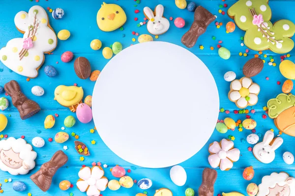 Easter sweets and candies background. High-colored blue flat lay with various Easter chocolate eggs, treats, symbols of easter sheep, bunny, carrot, flowers, cookies, top view copy space, frame
