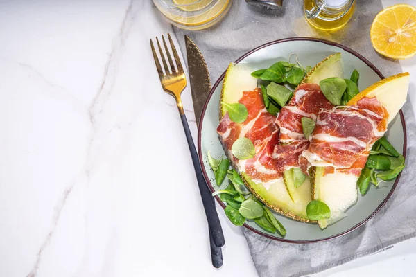 stock image Fresh melon salad with prosciutto hamon and green leaves mix, autumn fruit vegetable salad, portion on plate, on white marble background copy space