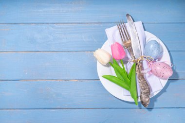 Easter table setting with tulip flowers and eggs, tag with text Happy Easter, over light blue wooden background top view copy space