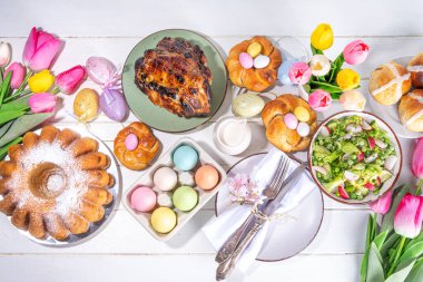 Festivalde Paskalya yemeği veya brunch masası sunuldu, Paskalya yumurtası, lale çiçeği buketi, geleneksel Paskalya yemekleri - pasta, çapraz çörekler, çelenk ekmeği, jöleli jambon ve bahar salatası, beyaz masa arkaplanı