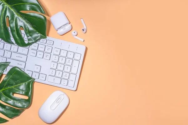 Summer office background with keyboard. Simple minimal beige flatlay with tropical monstera leaf, white keyboard, mouse, earphones, top view copy space
