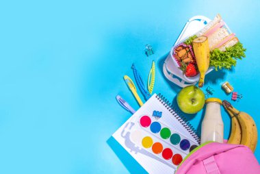 Sağlıklı okul beslenme çantası: sandviç, sebze, meyve, fındık ve yoğurt, içinde okul çocukları malzemeleri, aksesuarlar ve yüksek renkli mavi arka plan fotokopi alanı üzerine sırt çantası