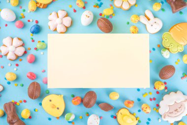 Easter sweets and candies background. High-colored blue flat lay with various Easter chocolate eggs, treats, symbols of easter sheep, bunny, carrot, flowers, cookies, top view copy space, frame