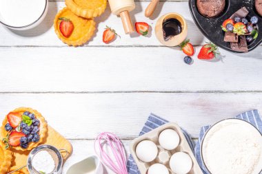 Fırında hamur işi ile yaz pişirme arka planı - çilekli kekler, mini turtalar, pişirme malzemeleri, un, yumurta, merdane, krema sütü, şeker, çilek, beyaz ahşap zemin