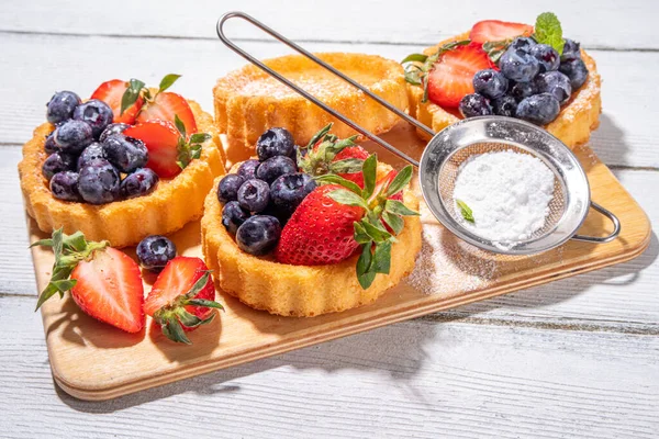 stock image Sponge mini tart cakes with summer berry. Portioned vanilla dessert cups with fresh strawberry, blueberry, raspberry, on white wooden background copy space