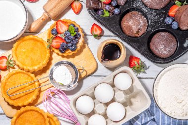 Fırında hamur işi ile yaz pişirme arka planı - çilekli kekler, mini turtalar, pişirme malzemeleri, un, yumurta, merdane, krema sütü, şeker, çilek, beyaz ahşap zemin