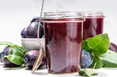 Red blue plum jam in small jar. Homemade autumn plum jam with fresh fruits. Fall preparations and canning on white table background copy space