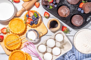 Fırında hamur işi ile yaz pişirme arka planı - çilekli kekler, mini turtalar, pişirme malzemeleri, un, yumurta, merdane, krema sütü, şeker, çilek, beyaz ahşap zemin