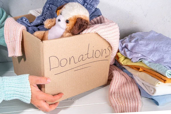 Donation box with clothes, toys and food on white table background. Cardboard donation box full with toys, female and child clothes, foods. Social care, volunteering, charity concept copy space