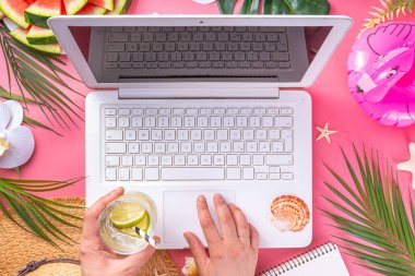 Yazlık ofis, çalışma geçmişi, blogcu iş yeri düzlüğü. Not defteri, hasır şapka, tropikal dekor ve yapraklarla dolu beyaz dizüstü bilgisayar klavyesi, açık pembe arka planda soğuk kokteyl bardağı.