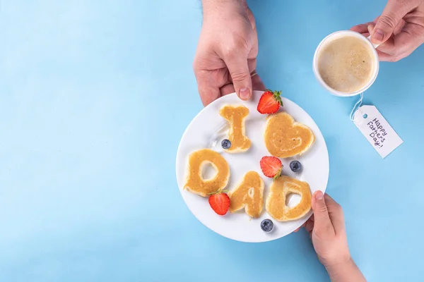 Father\'s day holiday idea for a cute surprise gift. Child preparing breakfast for dad, creatively decorated pancakes with the inscription I love Dad. Top view copy space, blue background