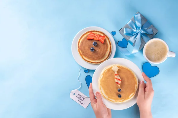 Father\'s day holiday idea, cute surprise gift. Child preparing breakfast for dad, creative decorated pancakes with tie, bow tie, inscription I love Dad. On light blue background, with child, dad hands