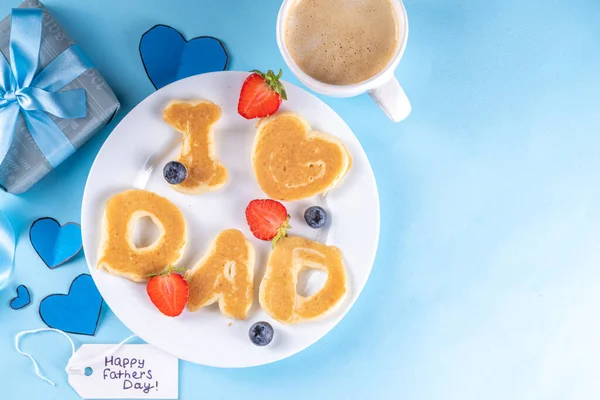 Father's day holiday idea for a cute surprise gift. Child preparing breakfast for dad, creatively decorated pancakes with the inscription I love Dad. Top view copy space, blue background