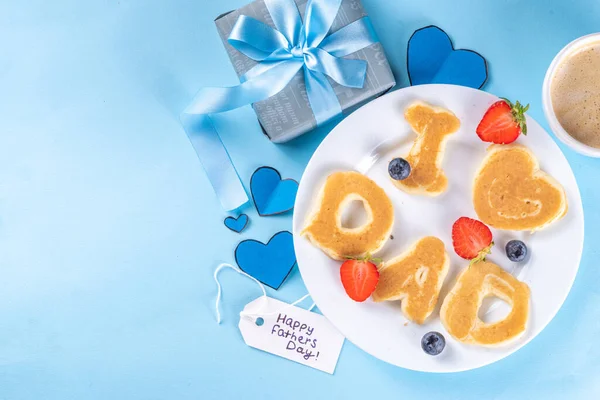 Father's day holiday idea for a cute surprise gift. Child preparing breakfast for dad, creatively decorated pancakes with the inscription I love Dad. Top view copy space, blue background