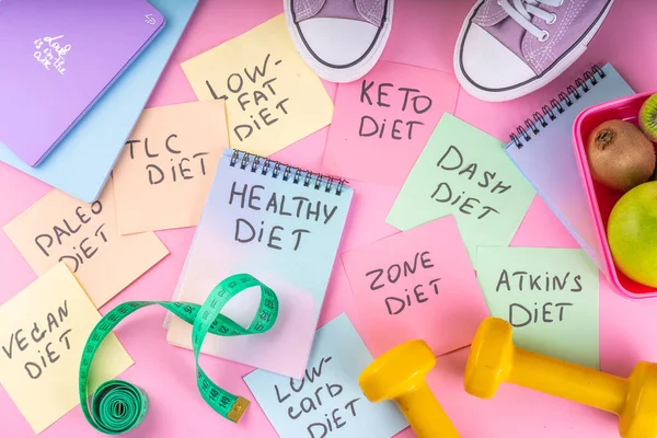 Slimming, fitness flat lay. Diet choice concept with bright handwritten notes with names of trendy diets and healthy eating concept, with sneakers, tape measure, dumbbells on bright pink background.