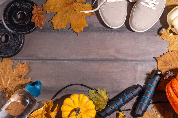 Autumn sport fitness background. Outdoors fall workouts concept. Running sneakers, dumbbell barbell discs, jump rope, water bottle, wooden background, with autumn leaves, pumpkins top view copy space