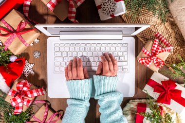 Tahta arka planda Noel hediyesi kutuları olan dizüstü bilgisayar kullanan biri. Kadın eli hediyeli defter, köknar ağacı ve kış dekoru. Noel ve Yeni Yıl için Hazırlık