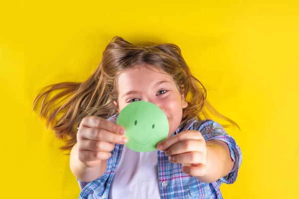 Mental health day, Protecting children from stress concept. Positive thinking, childhood depression therapy. Cute european pre-teenage girl holds paper smiley emoticon in her hand on yellow background