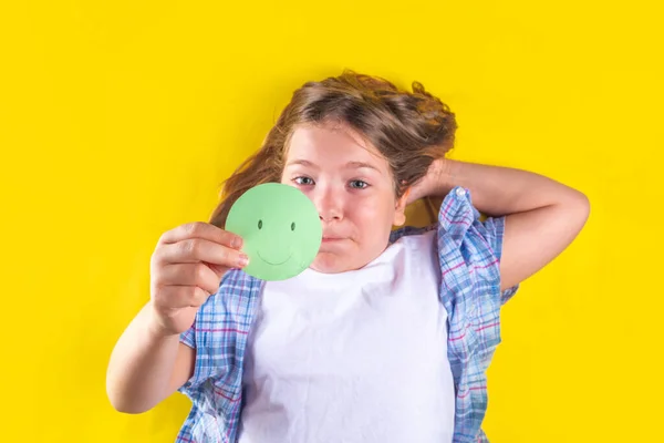 Mental health day, Protecting children from stress concept. Positive thinking, childhood depression therapy. Cute european pre-teenage girl holds paper smiley emoticon in her hand on yellow background