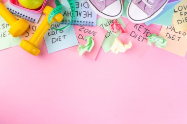 Slimming, fitness flat lay. Diet choice concept with bright handwritten notes with names of trendy diets and healthy eating concept, with sneakers, tape measure, dumbbells on bright pink background.