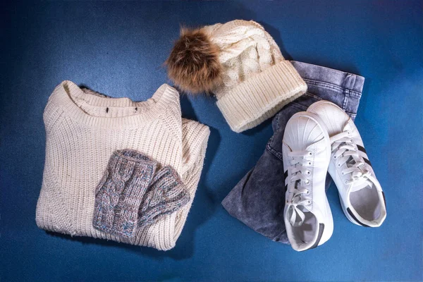 Set of female winter autumn clothes. Jeans, white knitted sweater and hat,  mittens and sneakers on  dark blue background. Casual cozy fashion outfit flat lay