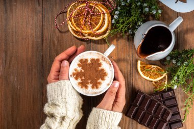 Cappuccino, ılık ahşap zemin üzerinde kar tanesi desenli sıcak çikolatalı latte kahve, kadın ellerinde sıcak çikolata içeceği olan fincan, üst görünüm fotokopi alanı. Noel sıcacık kafeterya afişi