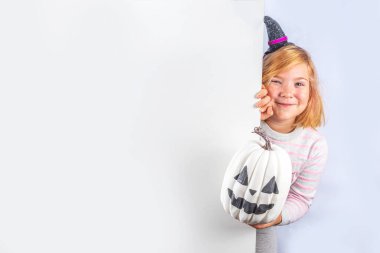 Happy Halloween holiday greeting card background. Happy cute european blonde girl in striped sweater with witch costume hat and jack carved decor pumpkin on white background copy space for text
