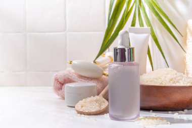 Pirinç suyu. Doğal organik pirinç kozmetiği. Mayalanmış güzellik, sağlık ve kaplıca. Şeffaf beyaz serum, beyaz banyo arka planında pirinç taneleri ve banyo malzemeleriyle şişe düşürür.