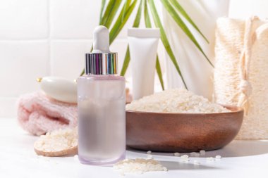Pirinç suyu. Doğal organik pirinç kozmetiği. Mayalanmış güzellik, sağlık ve kaplıca. Şeffaf beyaz serum, beyaz banyo arka planında pirinç taneleri ve banyo malzemeleriyle şişe düşürür.