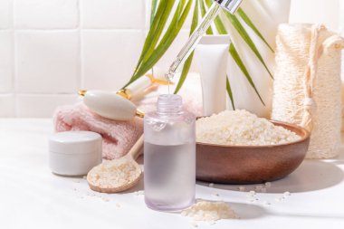 Pirinç suyu. Doğal organik pirinç kozmetiği. Mayalanmış güzellik, sağlık ve kaplıca. Şeffaf beyaz serum, beyaz banyo arka planında pirinç taneleri ve banyo malzemeleriyle şişe düşürür.
