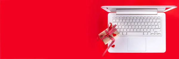 Valentine day simple background, Woman using laptop, with gift bow with festive tied ribbon, heart decor, on bright red background. Online Valentine`s shopping, preparation for February 14 holiday.
