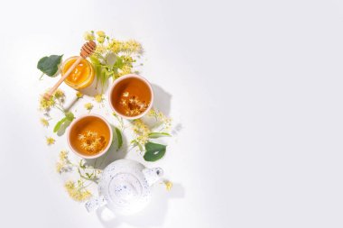Organik bitkisel sağlık içeceği. Linden çiçeği yeşili çay, beyaz kupalar, çaydanlık, beyaz bir masa, kopya alanı ve çiçek açan ıhlamur çiçekleri