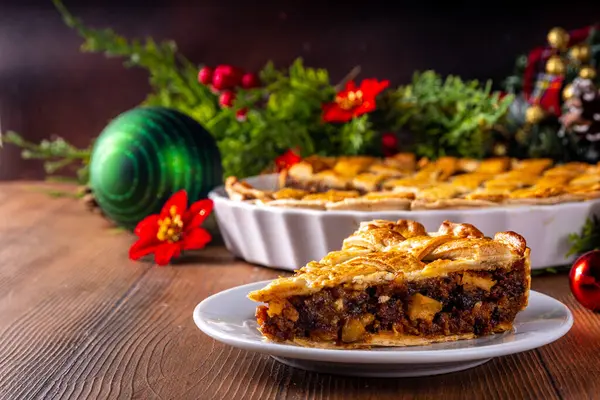 Traditional British Christmas mince pie. Homemade sweet mincemeat tart cake, filled with spiced dried fruit, nuts and apples, on cozy wooden table with  with Christmas tree and decoration