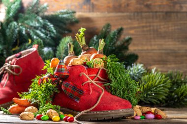 Sinterklaas, Saint Nicholas day background. Little kid red shoe with carrots and sweets, trooigoed, pepernoten, candy. Dutch Christmas holiday on cozy wooden background. 