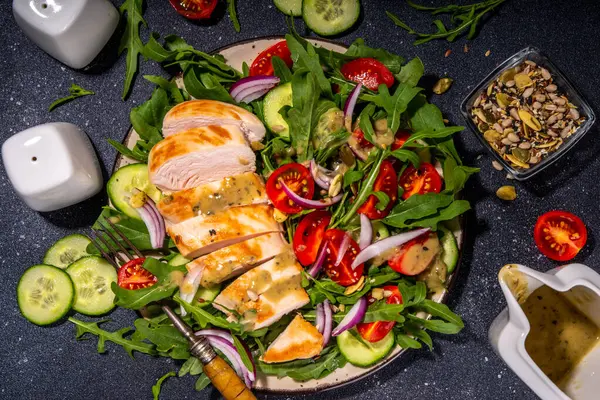 Grilled chicken fillet with vegetable salad. White meat, chicken or turkey breast, with fresh vegetables and greens salad, tomato, arugula, cucumber, onion, green olive oil sauce on black background