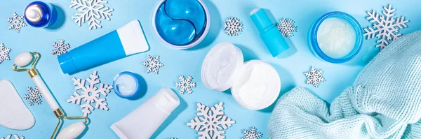 Winter skin care and hand care cosmetic in unbranded containers, bottles, tubes with artificial snowflakes on light blue background. Winter hand skin care cosmetics background top view copy space