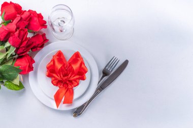 Valentine day table setting on white background. Romantic diner table setting for with red roses, heart shaped bow, plate, wine glass and cutlery top view copy space. Valentines menu background