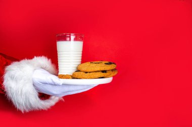 Noel Baba 'nın eli, geleneksel Noel kurabiyesi atıştırmalığıyla süt bardağıyla. Noel Baba Batıran Kurabiye, parlak kırmızı yırtık kağıt delikli arka planda. Mutlu Noeller ve mutlu yıllar tebrik kartı.