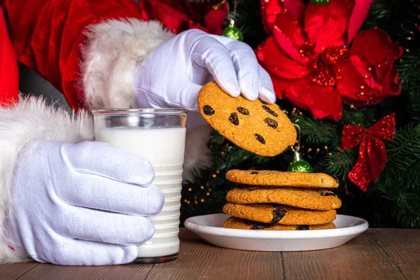 Santa Claus eating cookie and milk. Santa hands picking cookie and milk glass on wooden table against festive Christmas tree,