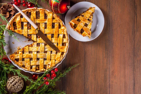 Traditional British Christmas mince pie. Homemade sweet mincemeat tart cake, filled with spiced dried fruit, nuts and apples, on cozy wooden table with  with Christmas tree and decoration